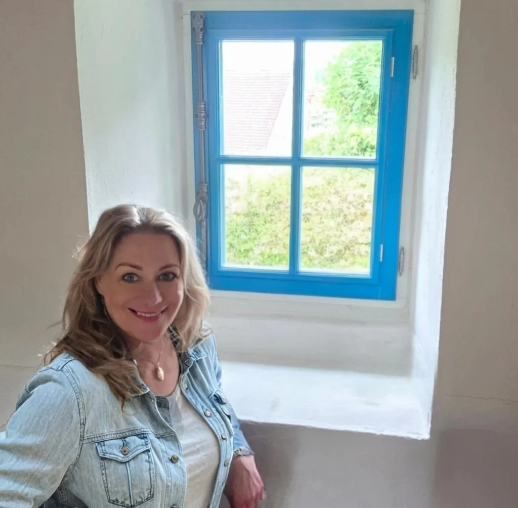 Marion staat voor een blauw raam, ze leunt op de vensterbank en kijkt vrolijk naar de camera.
