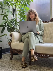 Een dame kijkt weg van de camer, ze zit op een antieke bank en heeft een laptop op haar schoot. ze en heeft lang bruin golvend haar, ze draagt een kaki pak. Ze staat tegen een halfbetegelde muur met verspatte