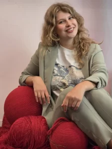 Een dame kijk met een schuin hoofd naar de camera, ze zit op een rode poef en heeft lang bruin golvend haar, ze draagt een kaki pak