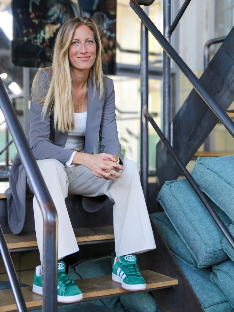 Irene, een dame met lang blond haar zit op een trap met stalen frame en houten tredes. Ze draagt groene gypmen, een kaki broek en een grijs colbert.