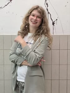 Een dame kijk met een schuin hoofd naar de camera, ze en heeft lang bruin golvend haar, ze draagt een kaki pak. Ze staat tegen een halfbetegelde muur met verspatten .