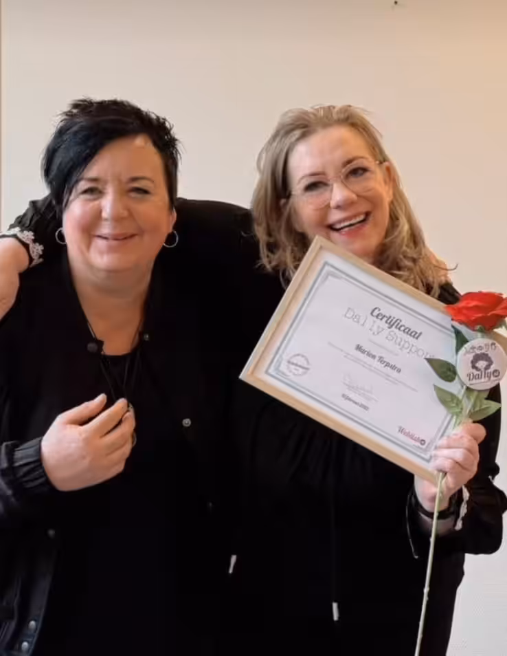 Een trotse Marion Terpstra toont haar behaalde 'Da11y Supporter' certificaat voor digitale toegankelijkheid. Ze staat lachend naast haar trainer met een rode roos in haar hand na de uitreiking.
