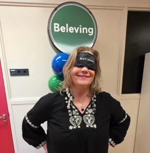 Marion Terpstra met een blinddoek op met de tekst 'Bartiméus', staand voor een bord met het woord 'Beleving'. Ze ervaart hier tijdens een training hoe het is om visueel beperkt te zijn, als onderdeel van haar expertise in digitale toegankelijkheid.