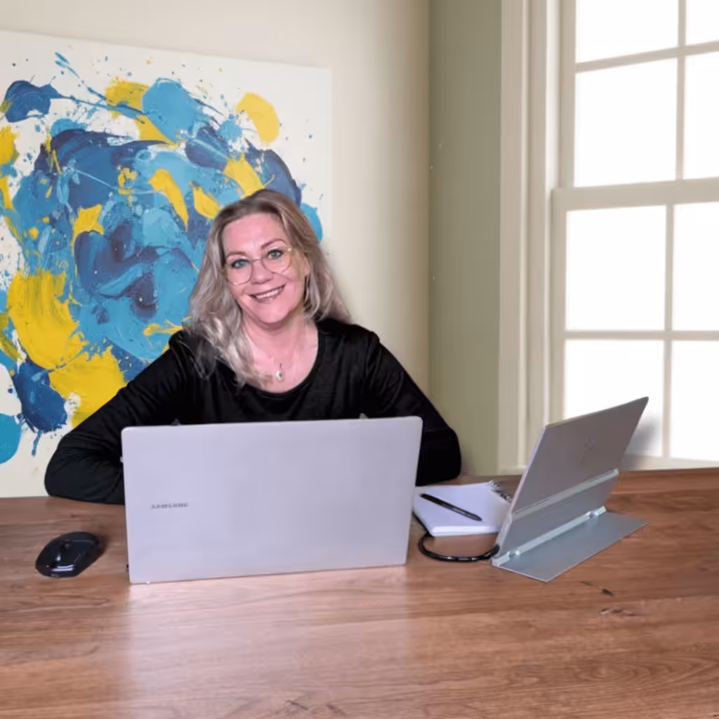 Marion Terpstra van Studio The WebCreator zit lachend achter haar laptop aan een houten bureau. Ze werkt aan een website strategie. Op de achtergrond hangt een groot kleurrijk abstract schilderij.