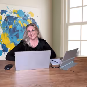 Marion Terpstra van Studio The WebCreator zit lachend achter haar laptop aan een houten bureau. Ze werkt aan een website strategie. Op de achtergrond hangt een groot kleurrijk abstract schilderij.