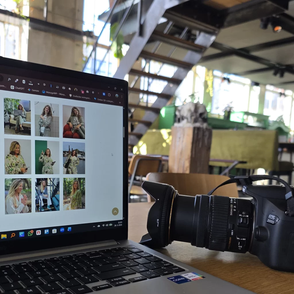 Een laptop staat open op een houten tafel en toont een raster van verschillende portretfoto's op het scherm. Rechts van de laptop ligt een zwarte Canon EOS 2000D spiegelreflexcamera met een grote Sigma lens. De achtergrond toont een sfeervolle, lichte kantooromgeving of café met een industriële trap en groene accenten.