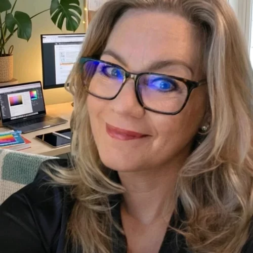 Een close-up van Marion, een vrouw met blond haar en een bril, die glimlachend in de camera kijkt. Op de achtergrond is haar werkplek zichtbaar met een laptop waarop kleurpaletten te zien zijn, een extra computerscherm, een tablet en een Monstera kamerplant. De blauwe weerspiegeling van het scherm is zichtbaar in haar glazen, wat de focus op haar digitale werk als ontwerper benadrukt.